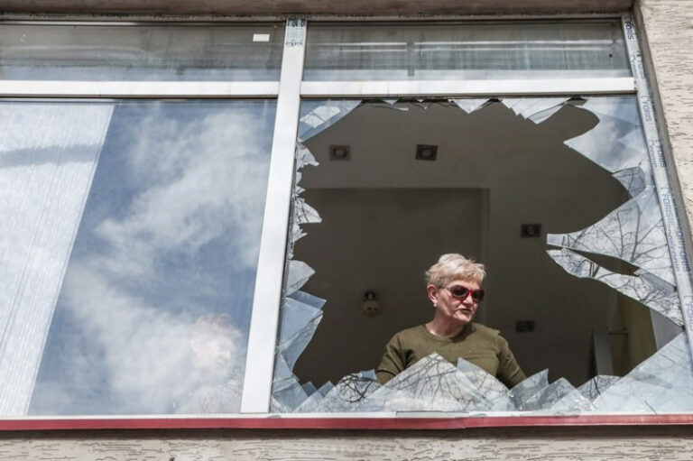 Жінка дивиться крізь розбите вікно після ворожих обстрілів міста. Фото: Stas Yurchenko для «Ґрати»