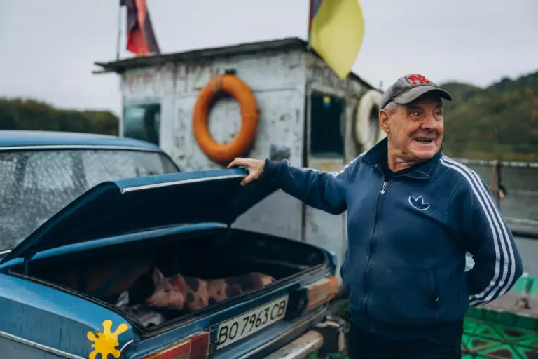 Чоловік перевозить на поромі через Дністер порося, розмістивши його в багажнику свого авто. На межі Буковини й Поділля. Жовтень 2025 року. Фото: Андрій Якименко.
